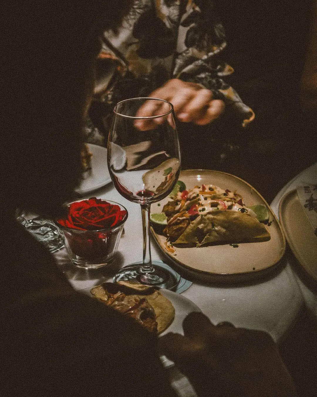 Intimate dining setting with a glass of wine, a lit red rose candle, and a plate of tacos